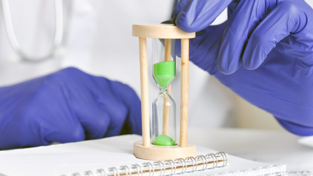 A doctor in medical gloves holds an hourglass.